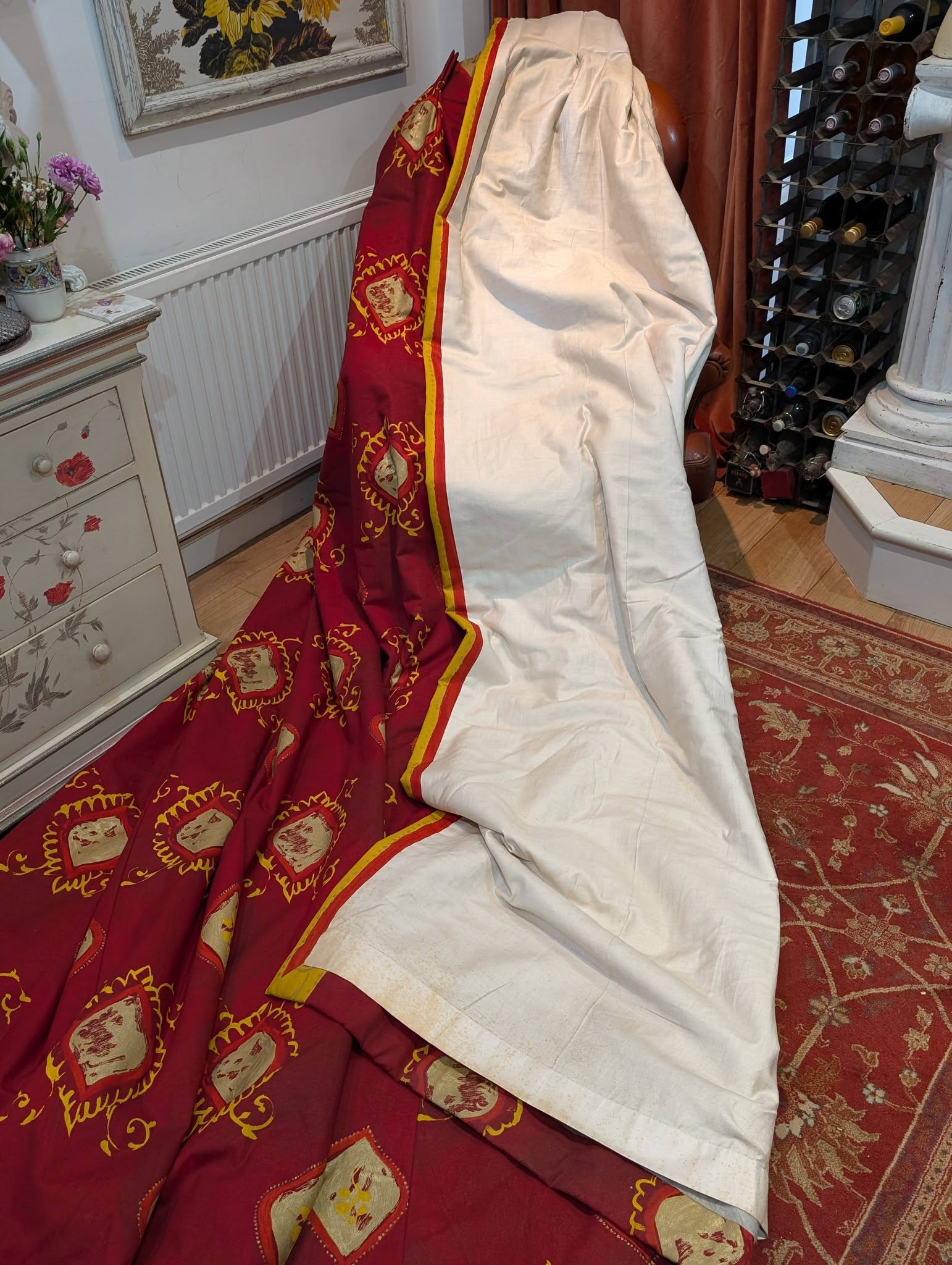 lauren7 - Divinely Vintage Interiors A tall object covered with white fabric beside a red and gold patterned cloth in a cozy room with a wine rack nearby.
