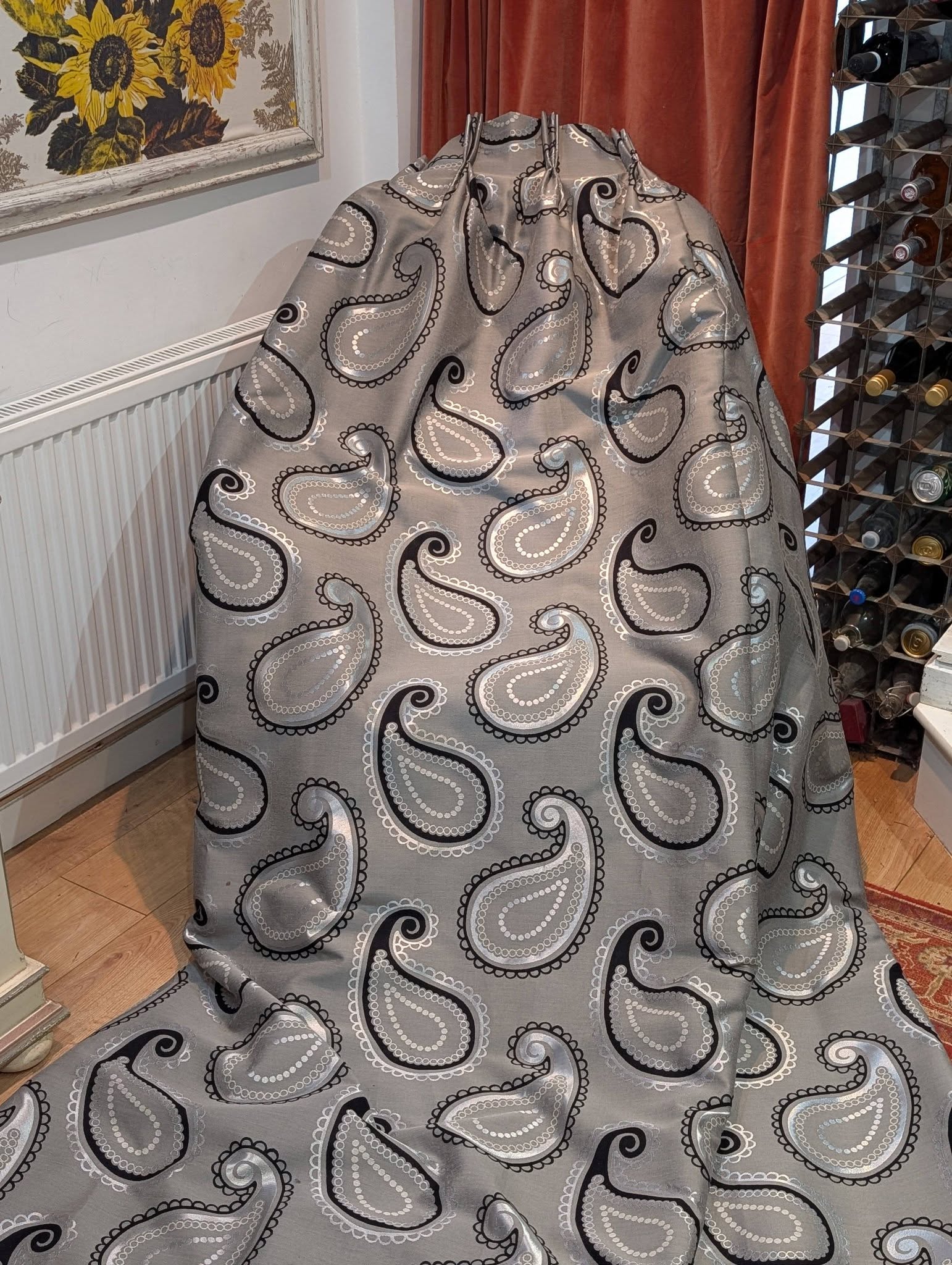 rach13 - Divinely Vintage Interiors Grey fabric with a white-and-black paisley pattern draped over a rounded object in a living room corner, with orange curtains nearby and a sunflower picture on the wall.