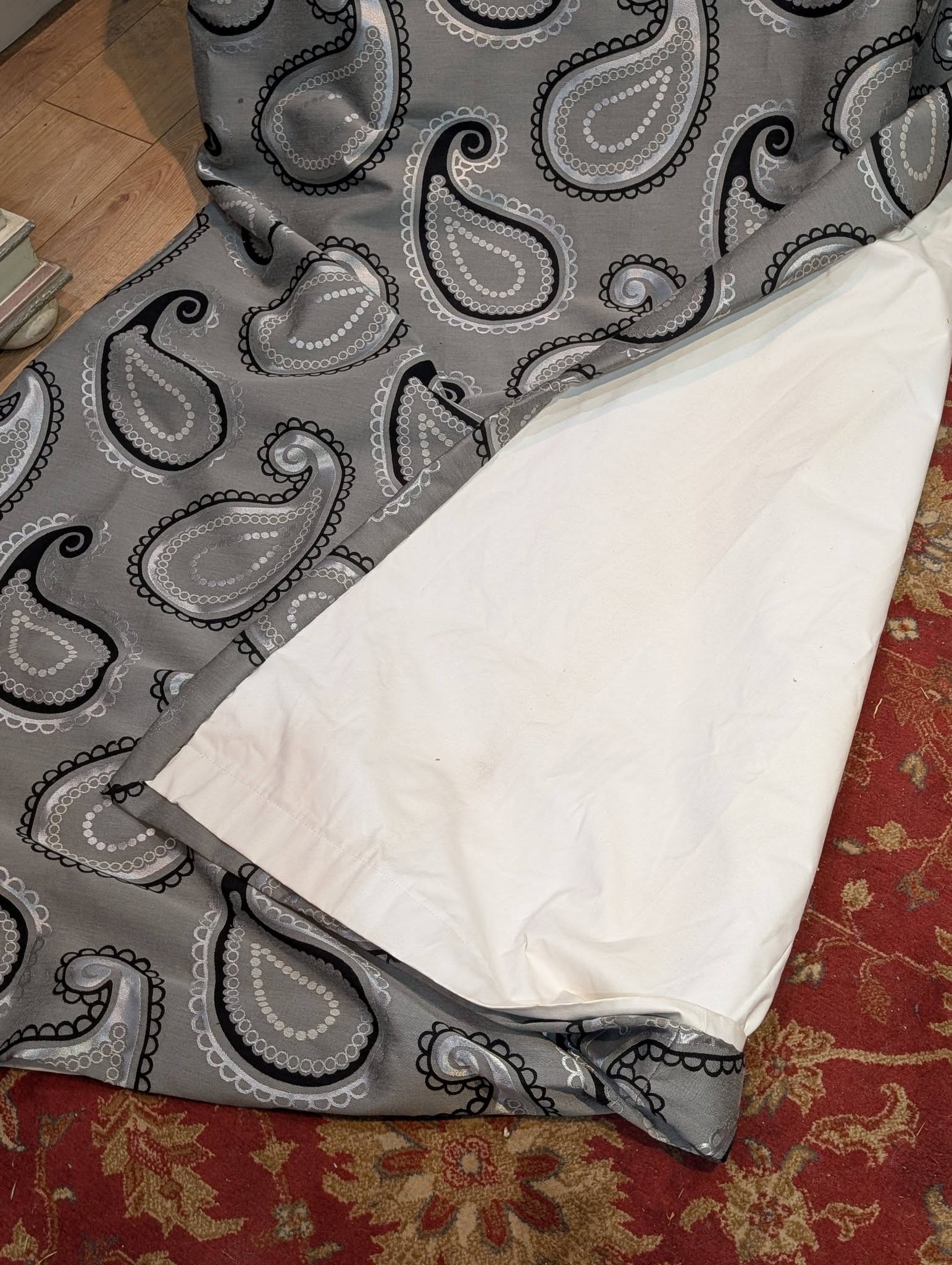 rach14 - Divinely Vintage Interiors Grey paisley-patterned fabric draped over a surface, with a white sheet and a red patterned carpet visible nearby.