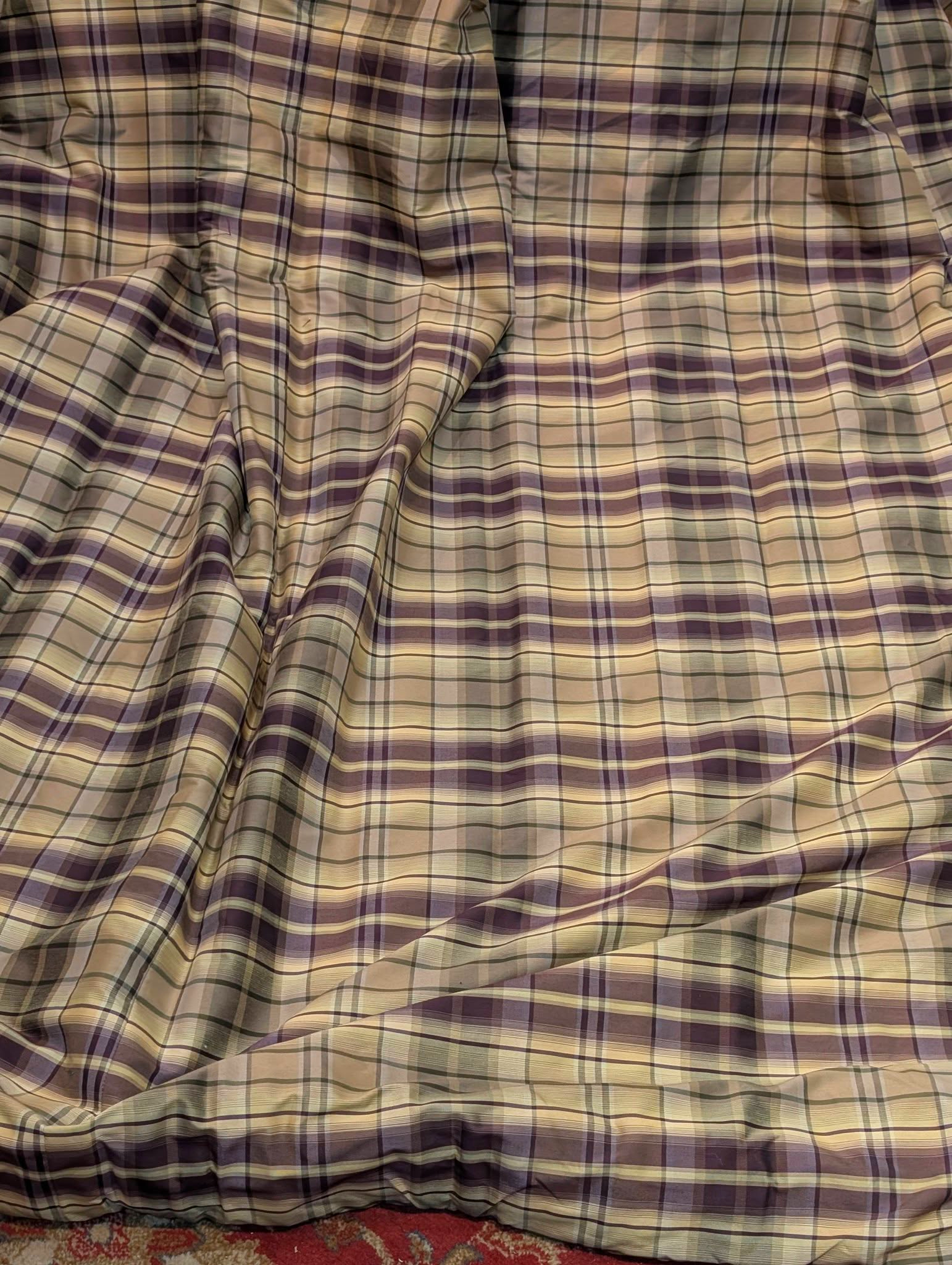 rach2 - Divinely Vintage Interiors Wrinkled plaid bedspread in purple, beige, and gray draped over a bed, with a red carpet edge visible at the bottom.