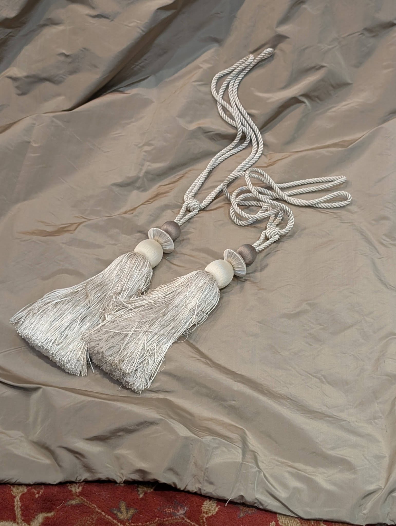 rach22 - Divinely Vintage Interiors Light gray tassel garland with wooden beads laid on a pale gray draped fabric backdrop.