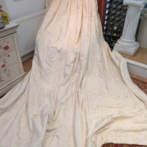 Large cream-colored quilted fabric draped over an unseen object in a living room, forming a rounded, tent-like shape with fabric pooling on the floor; background shows a radiator, floral painting, red curtains, a wine rack, and a white column.