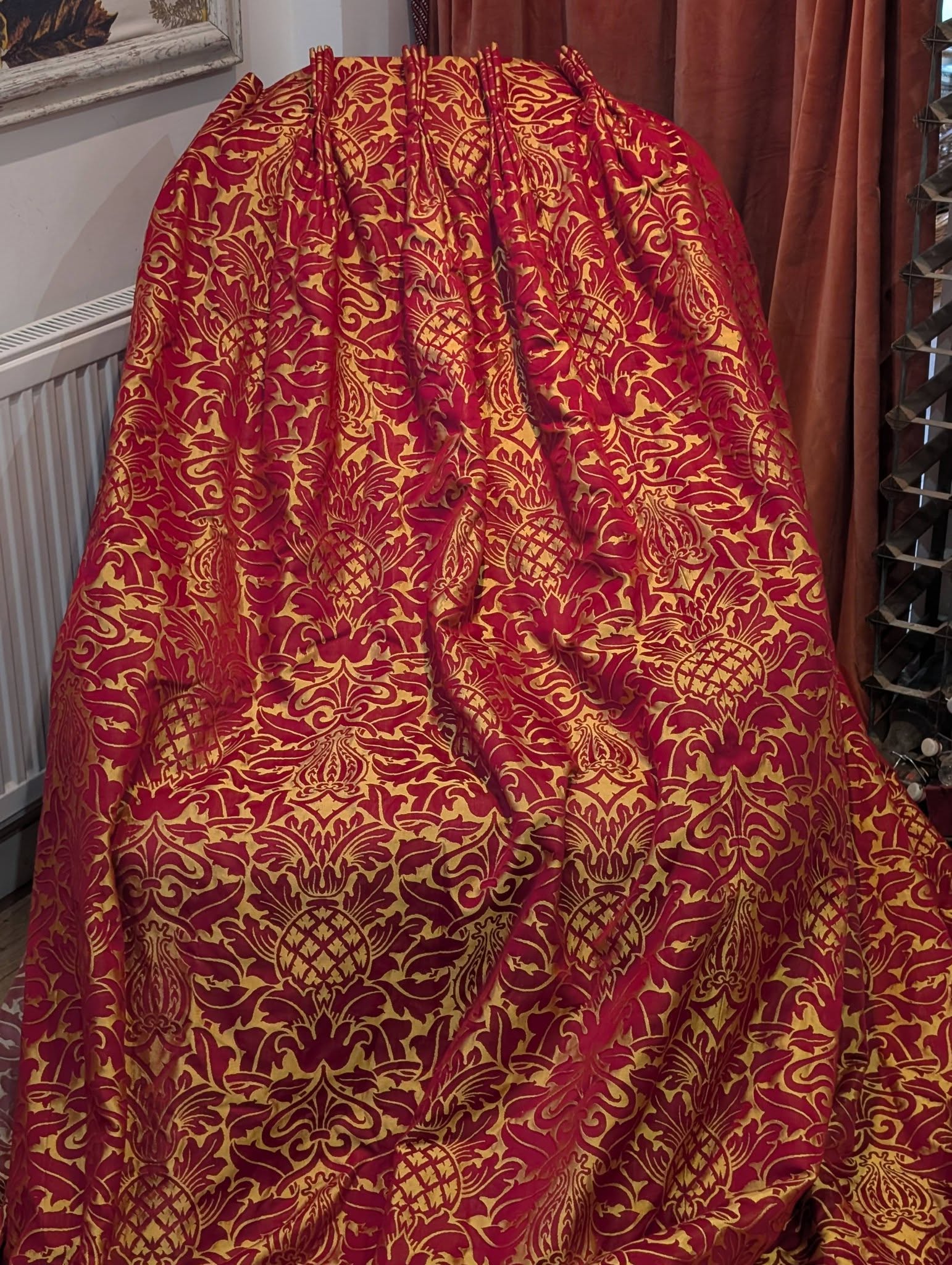 rich12 - Divinely Vintage Interiors Red and gold damask fabric draped over a stand in a fabric shop, showing an ornate floral pattern.