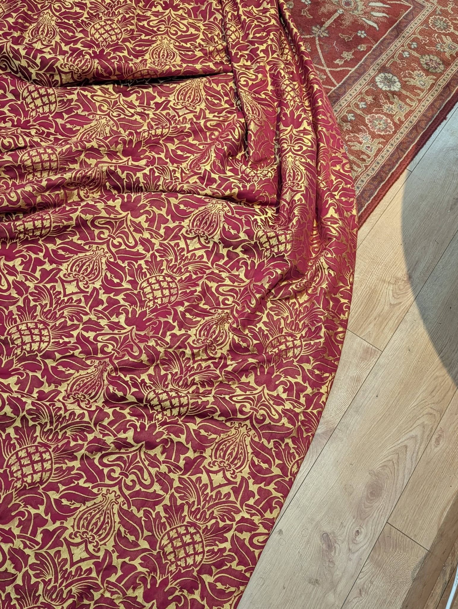rich16 - Divinely Vintage Interiors Red and gold damask-patterned fabric laid on a light wooden floor, with a decorative rug edge nearby.