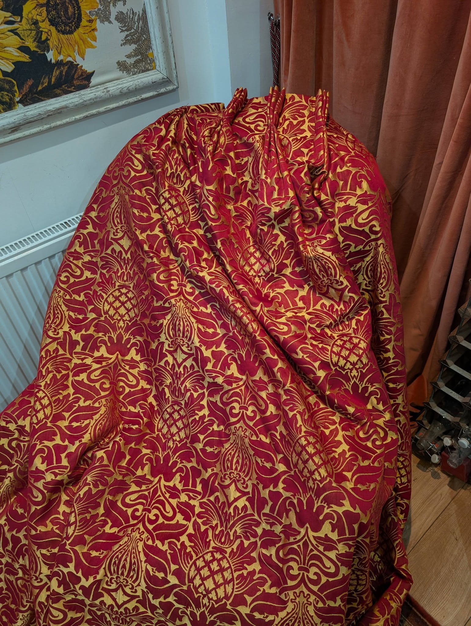 rich17 - Divinely Vintage Interiors Red and gold damask fabric draped over a chair or seating, with peach curtains and a sunflower-themed framed artwork in the background.