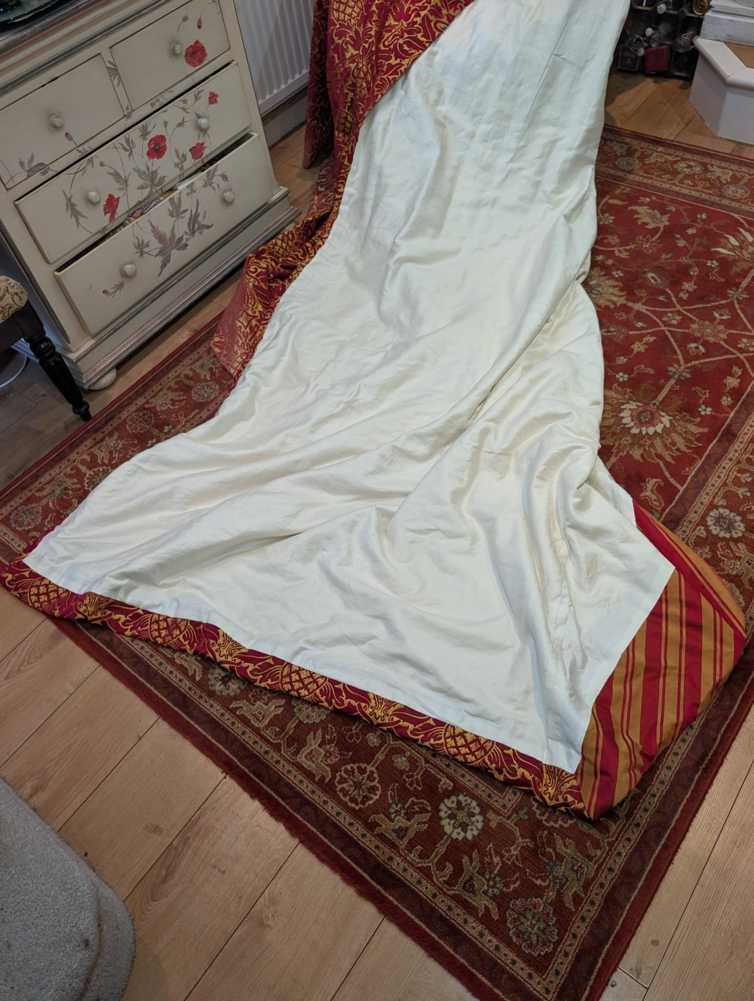 rich18 - Divinely Vintage Interiors A white bed sheet draped over a corner of a bed next to a floral painted dresser on a red patterned rug.