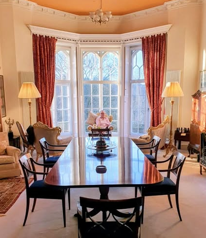 rich20 - Divinely Vintage Interiors Elegant dining room with a long glossy table, black chairs, and a person seated in a carved armchair by large bay windows with red drapes and warm lamps on either side.