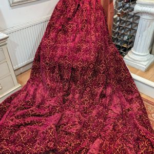 A large, magenta fabric with gold floral pattern drapes over an unseen chair or sculpture in a vintage room with a sunflower painting and wine rack nearby.