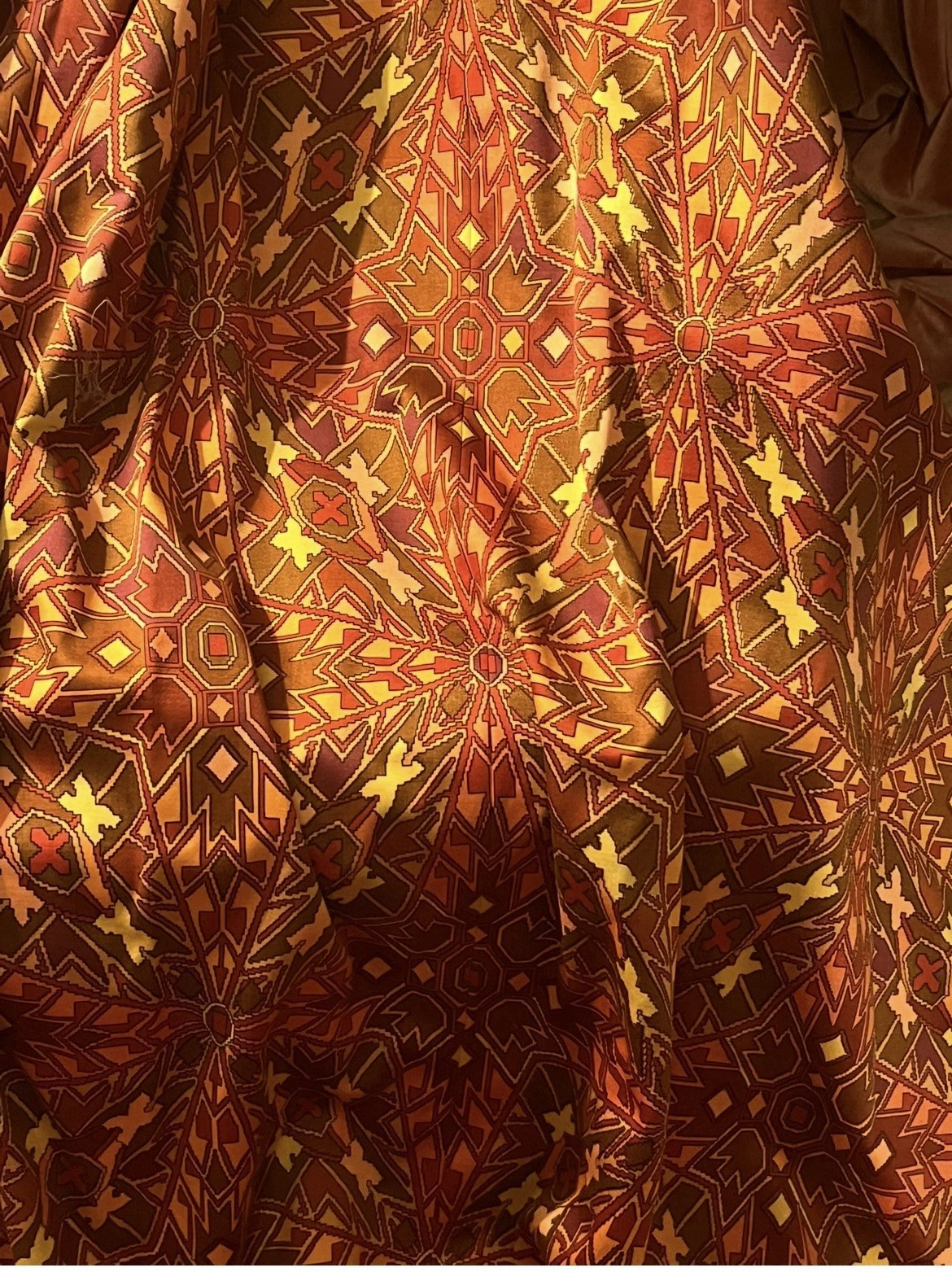akabah2 - Divinely Vintage Interiors Close-up of a patterned textile with geometric star-like motifs in red, orange, yellow against a brown background, with visible folds in the fabric.