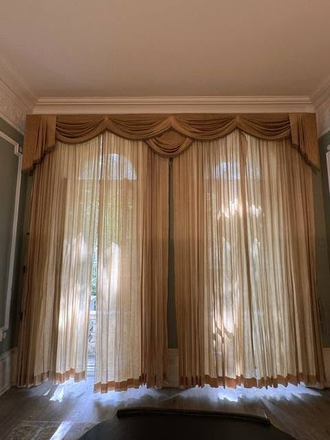 drew2 - Divinely Vintage Interiors Beige sheer curtains with a swag valance over tall arched windows, filtering sunlight into a formal living room.