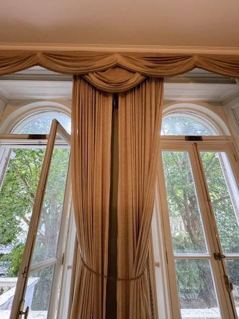 drew3 - Divinely Vintage Interiors Tall arched windows with beige drapes tied back, a swag valance at the top, letting in a leafy outdoor view.