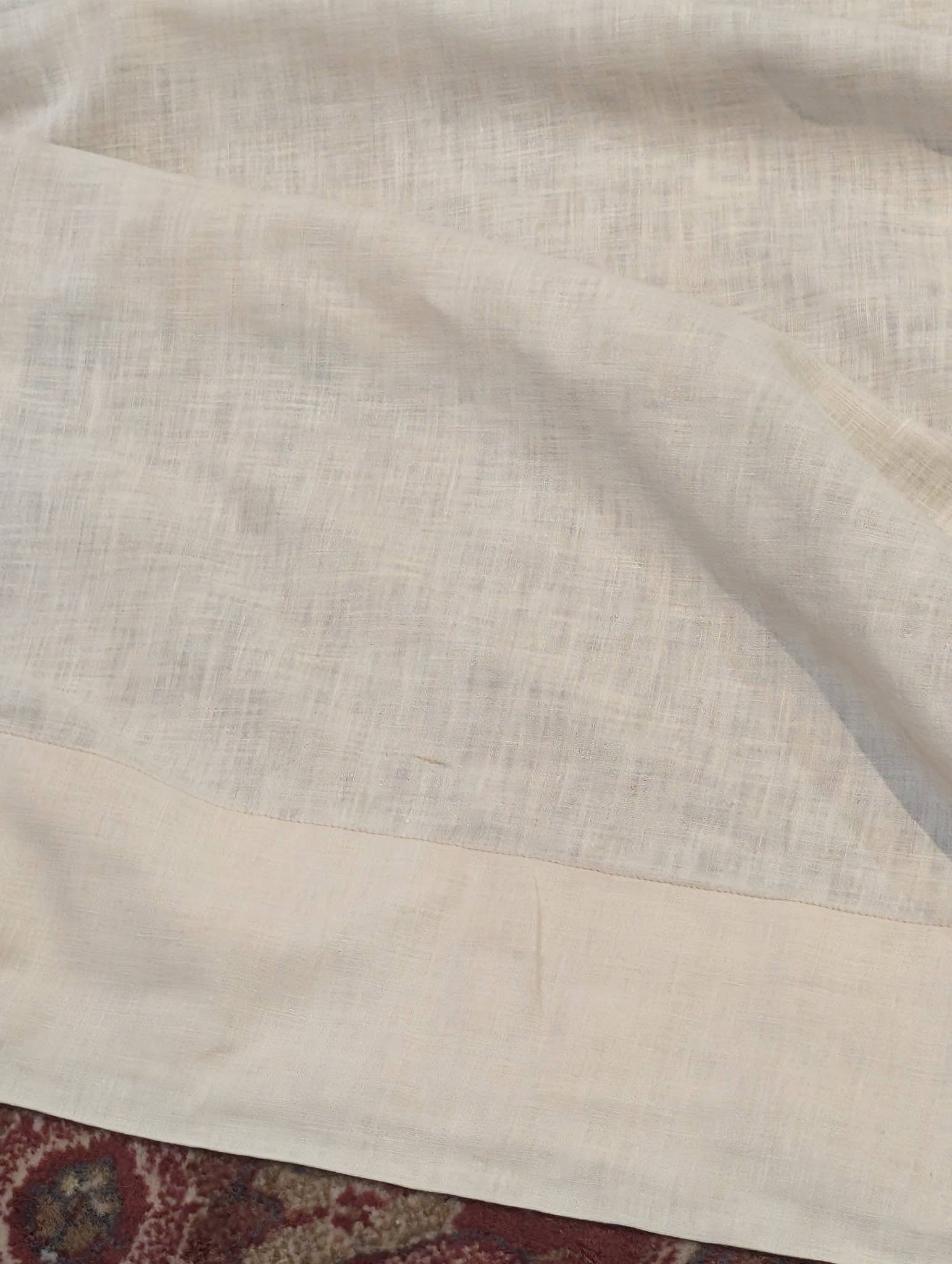 drew6 - Divinely Vintage Interiors Close-up of light beige textured fabric with a soft fold, resting over a surface with a rug edge visible at the bottom.