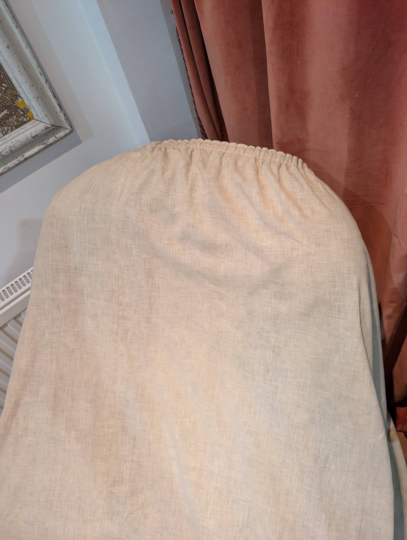 drew8 - Divinely Vintage Interiors Close-up of a beige fabric chair with a gathered edge, sitting in front of brown curtains and a white-framed window corner.