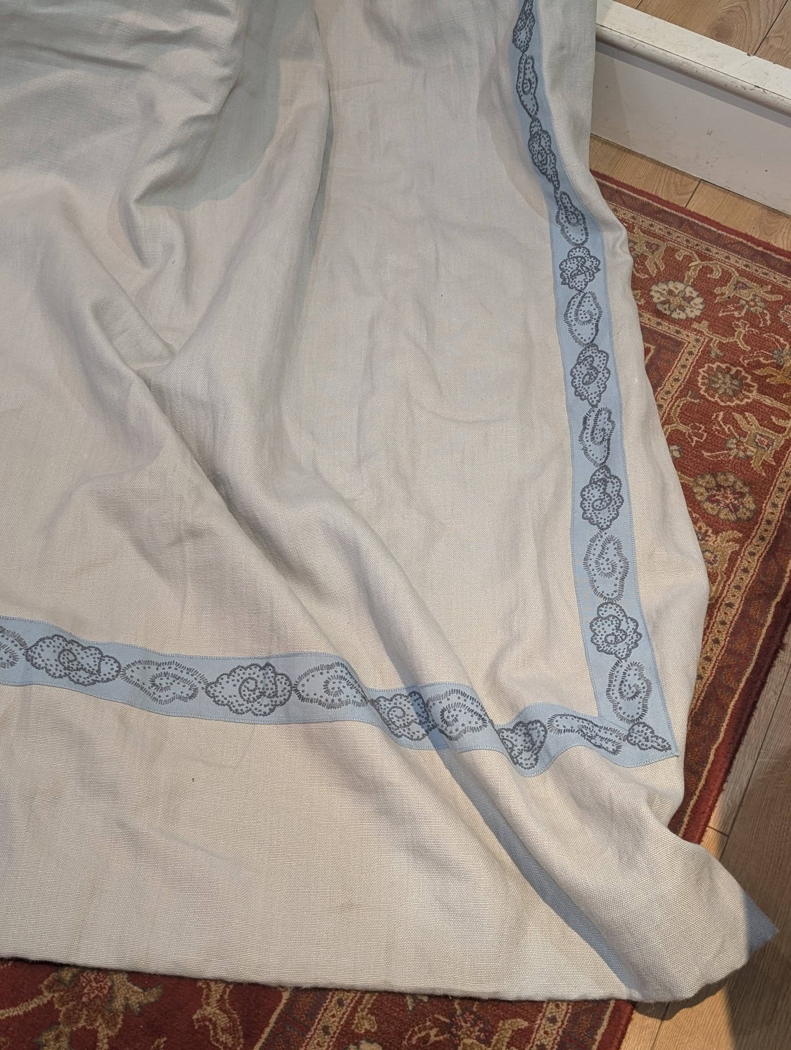 hannah13 - Divinely Vintage Interiors Light gray linen fabric with a pale blue embroidered border laid on a wooden floor and red rug.