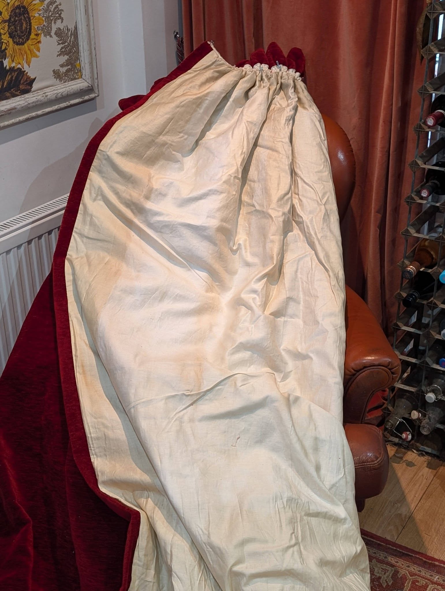 red7 - Divinely Vintage Interiors Cream-colored fabric with red trim draped over a burgundy chair in a living room, with a wine rack to the right and a sunflower picture on the wall.