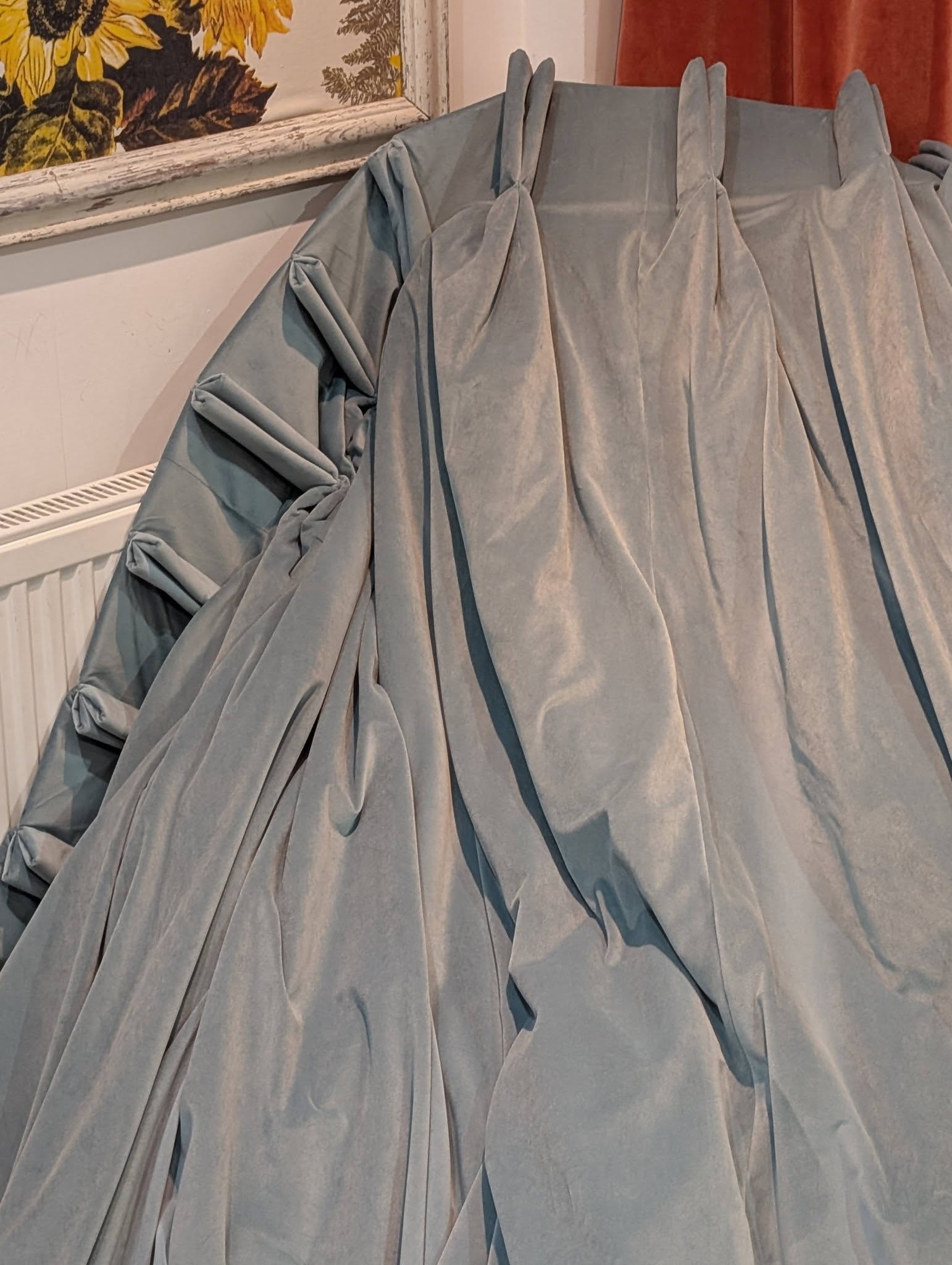 teal3 - Divinely Vintage Interiors Grey, heavy fabric draped into pleats and peaks in a corner of a room, with sunflower-patterned wallpaper and a radiator visible in the background.