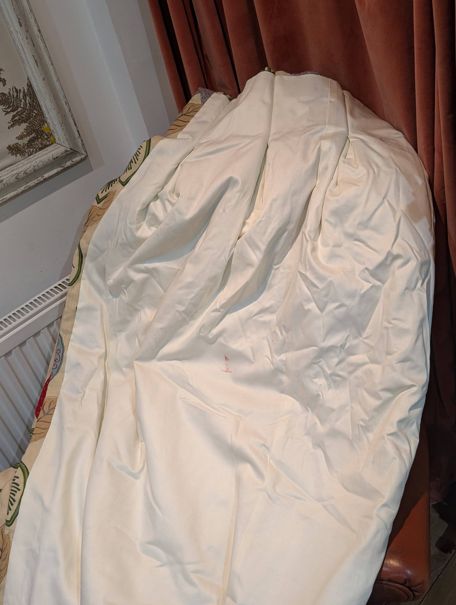 A pile of white bedding or fabric draped over an object beside a burgundy curtain, with a radiator vent visible on the left side.