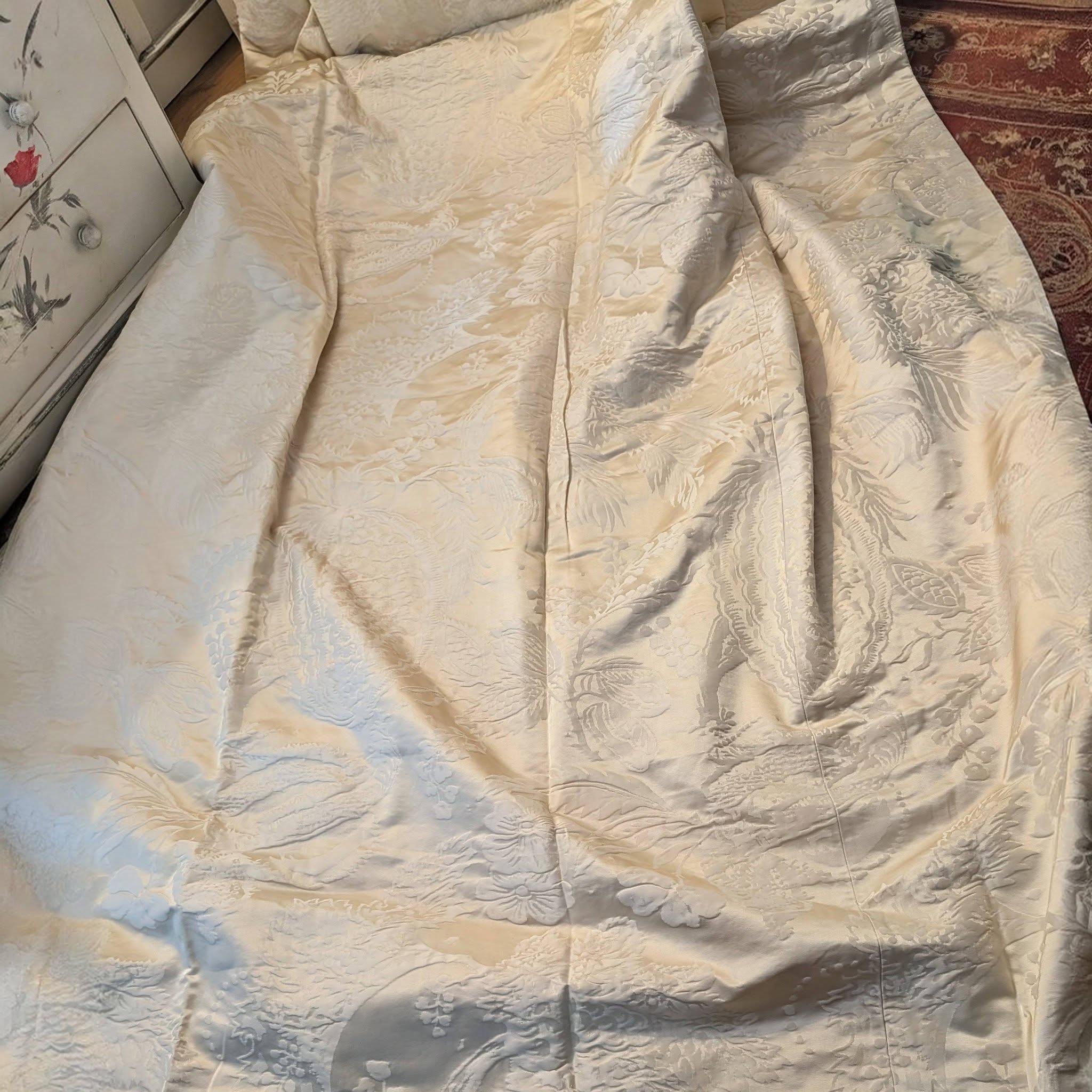 dam9 - Divinely Vintage Interiors Cream satin quilt with a subtle floral damask pattern spread across a bed next to a dresser and a red patterned rug nearby.