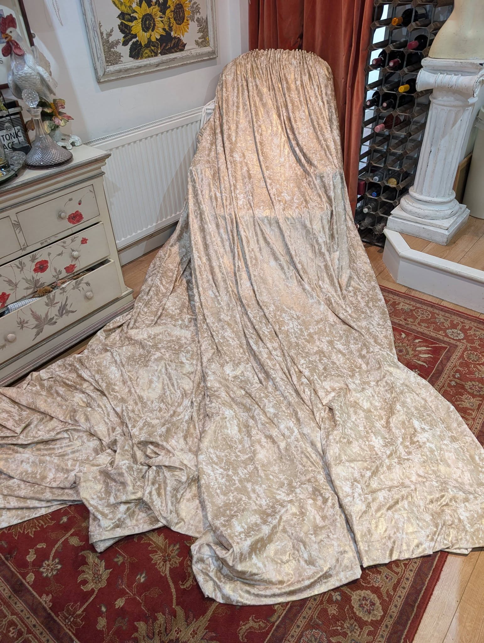 film1 - Divinely Vintage Interiors Chair covered in beige crushed-velvet fabric, pooling onto a red patterned rug in a vintage-style room with a sunflower painting and a wine rack nearby