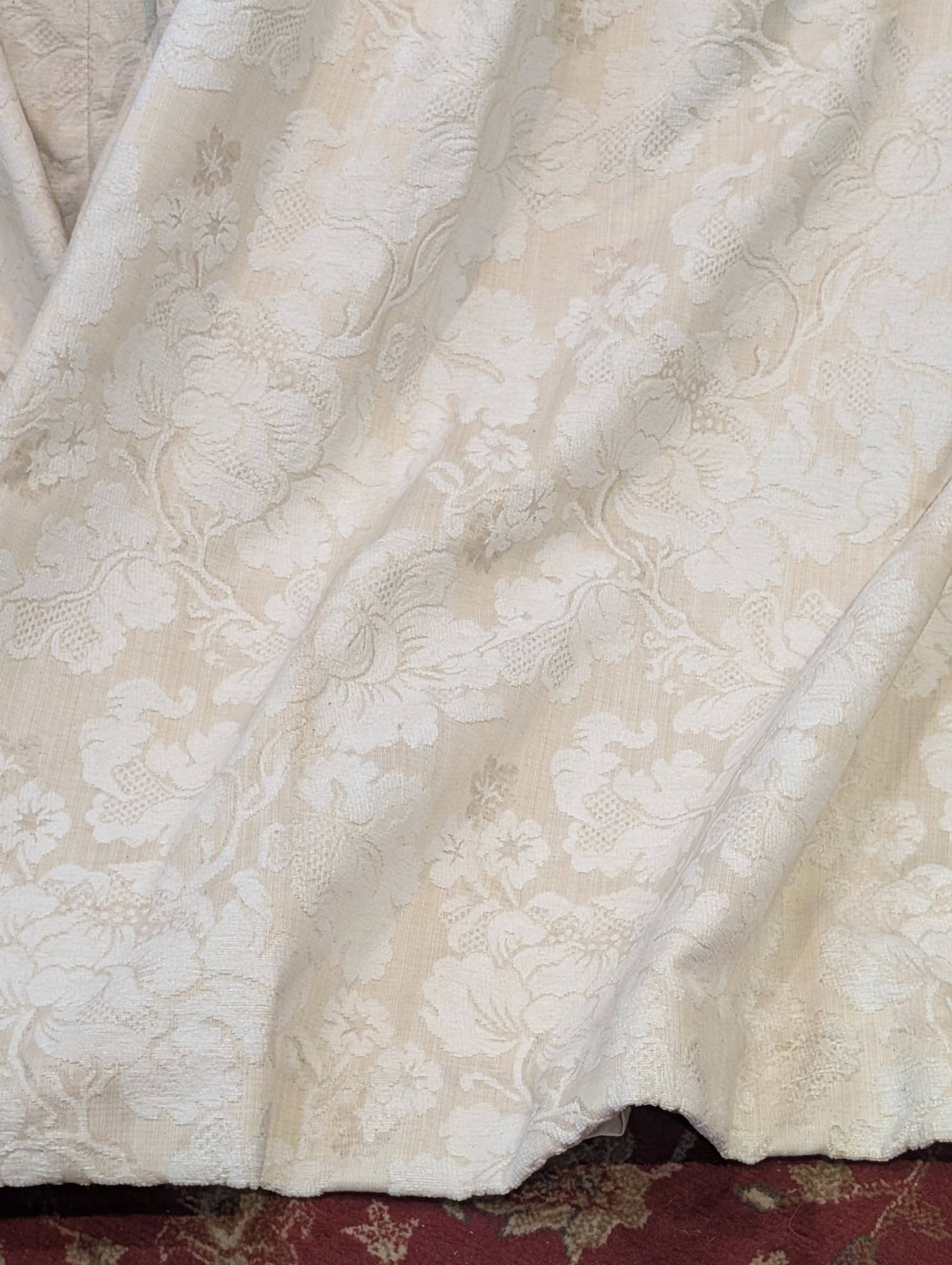 leila2 - Divinely Vintage Interiors Close-up of ivory lace fabric with floral pattern, part of a wedding dress draped over a dark red carpet edge.