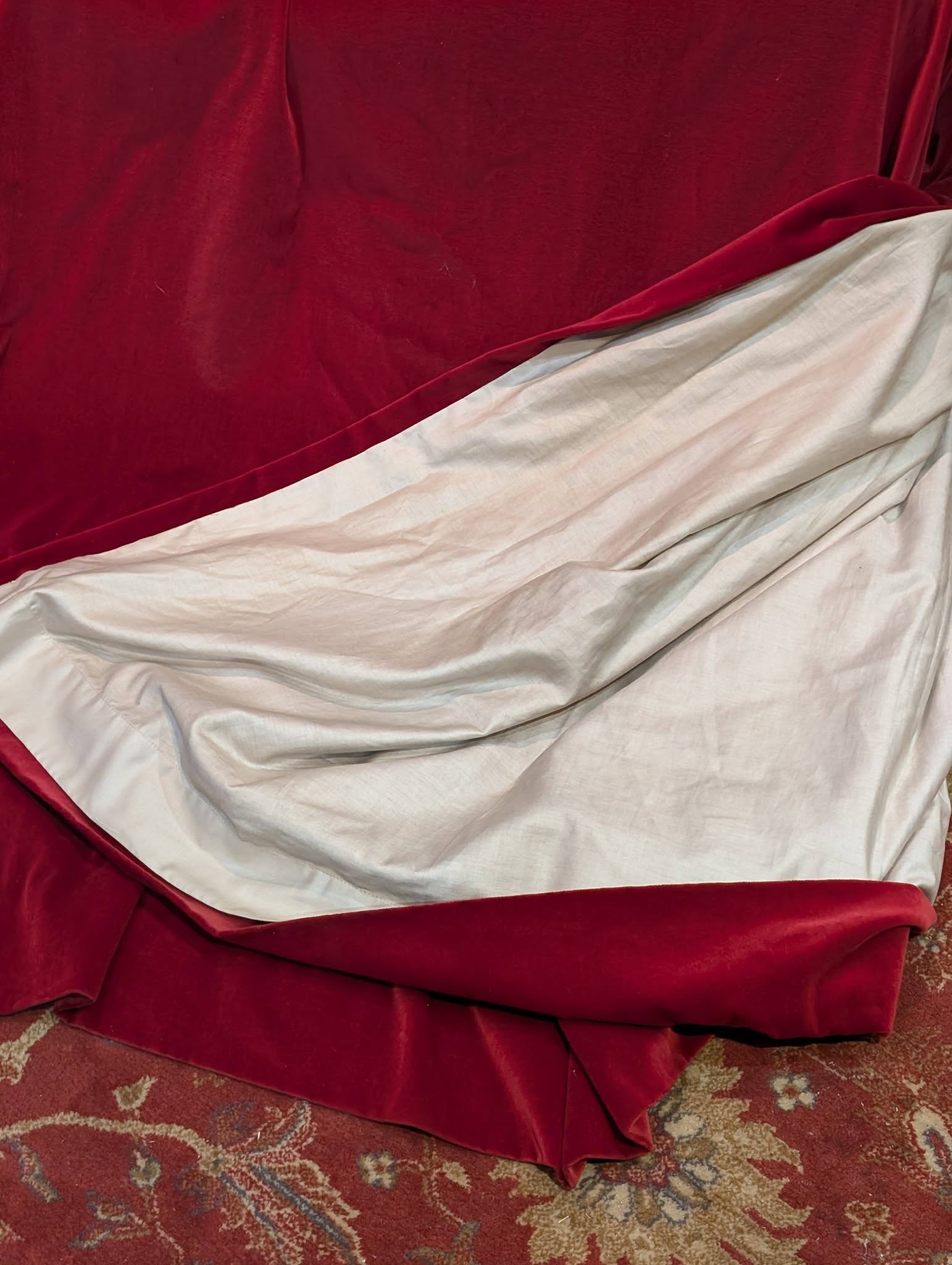 Red velvet blanket draped over a white sheet on a patterned rug, with folds creating soft shadows.