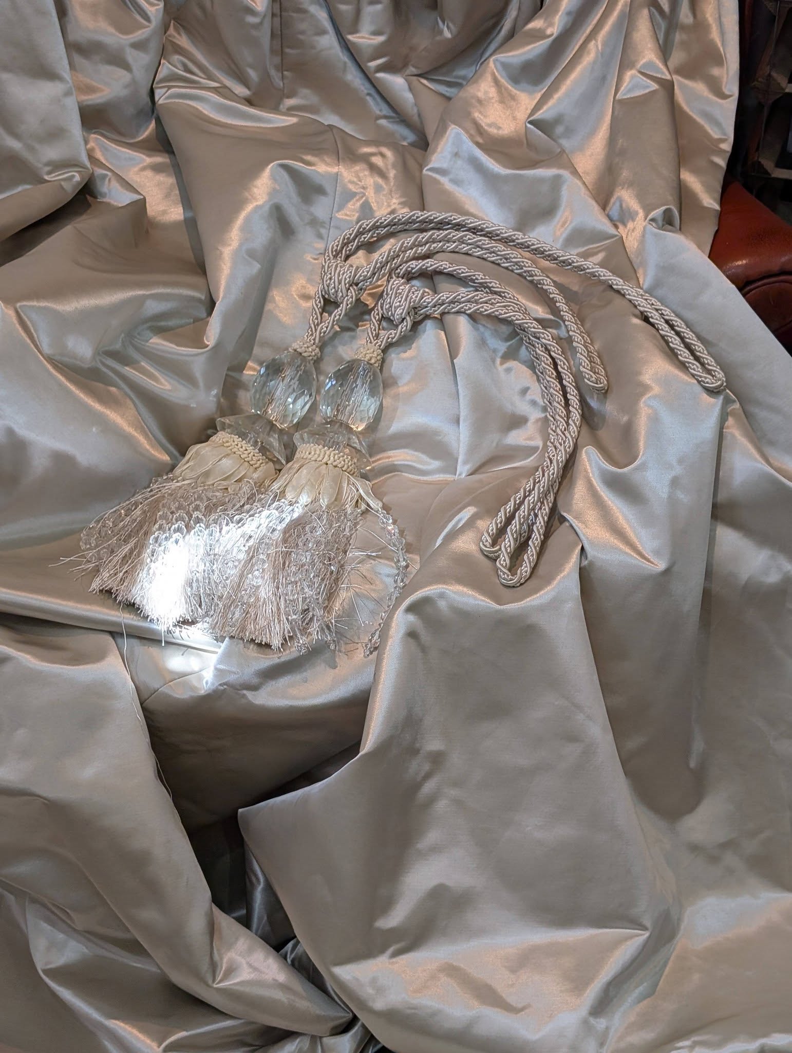 silk7 - Divinely Vintage Interiors Close-up of light gray satin fabric with twisted braided cords and crystal tassel ends laid on it.