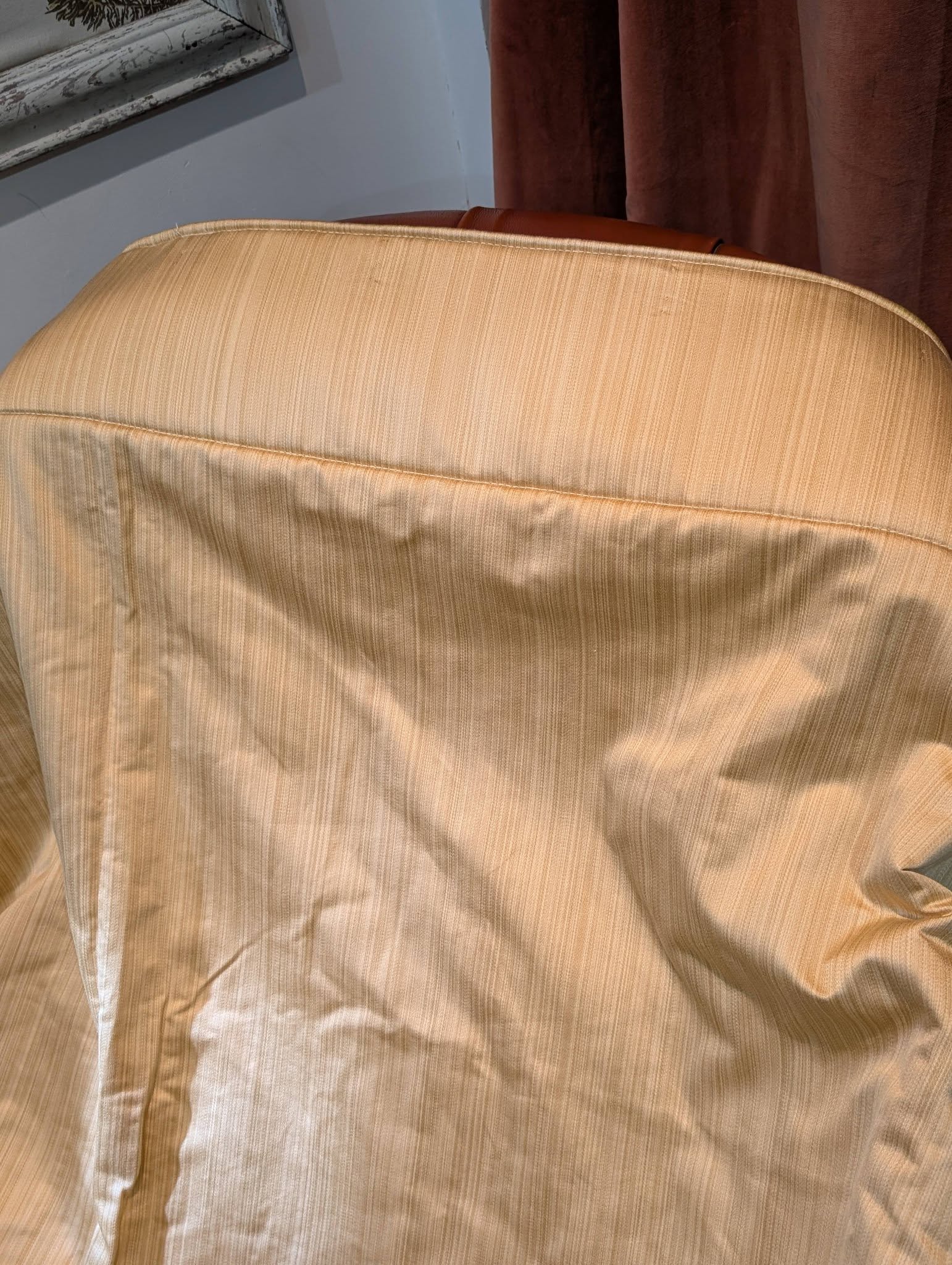 single5 - Divinely Vintage Interiors Close-up of a beige striped chair back in a room, with a window frame and brown curtains in the background.