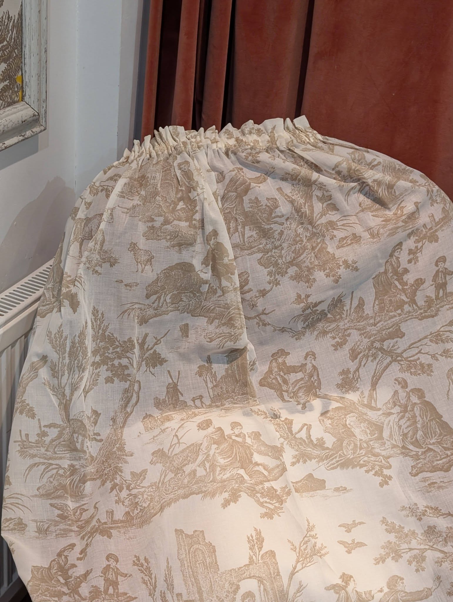 Beige toile-patterned fabric draped over a rounded piece of furniture in a room, showing pastoral scenes and figures.