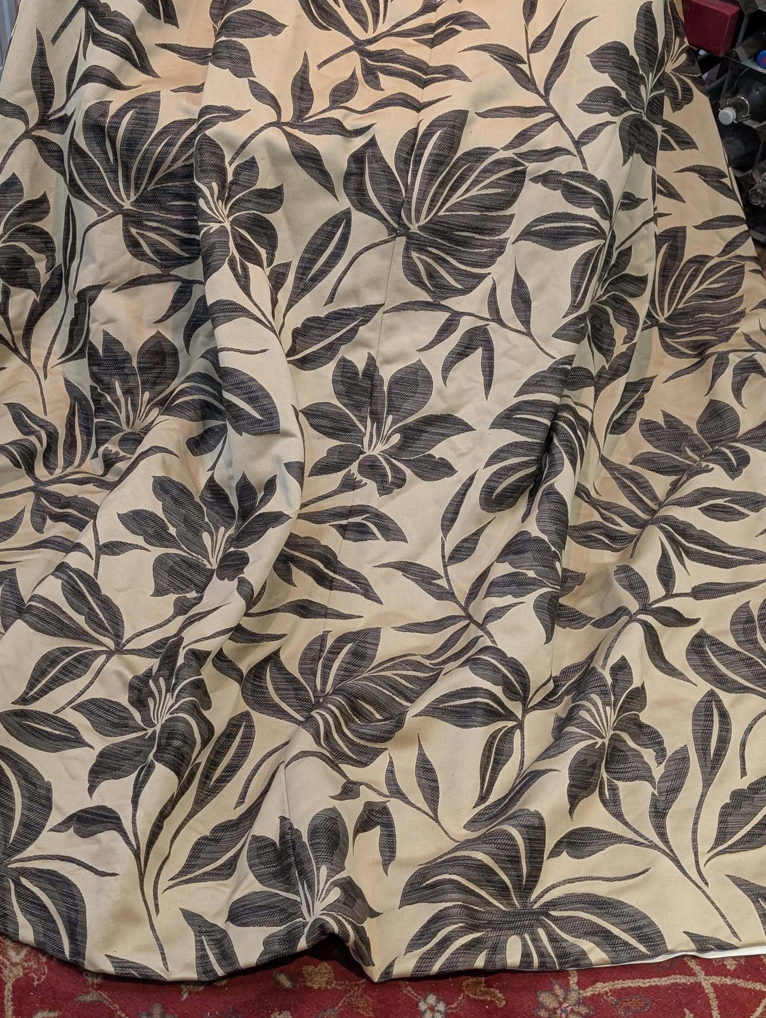 Beige fabric with a blue botanical leaf pattern and visible folds, lying on a red rug edge.