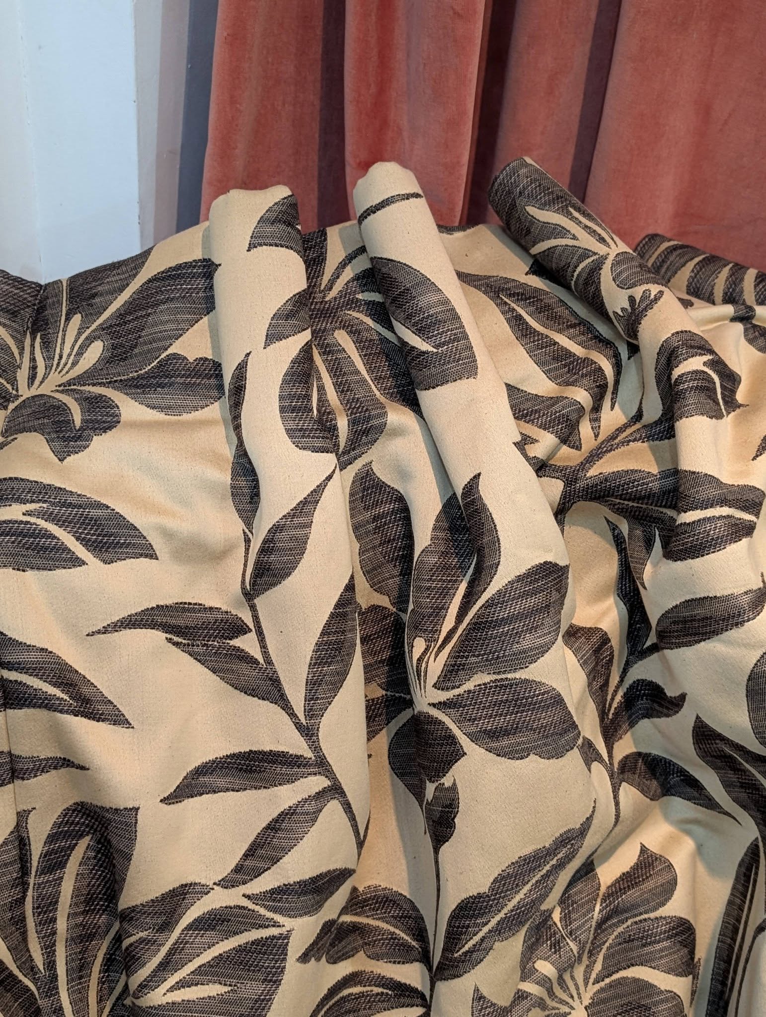 Close-up of beige fabric with a large dark botanical leaf print, draped over a surface with pink curtains in the background.